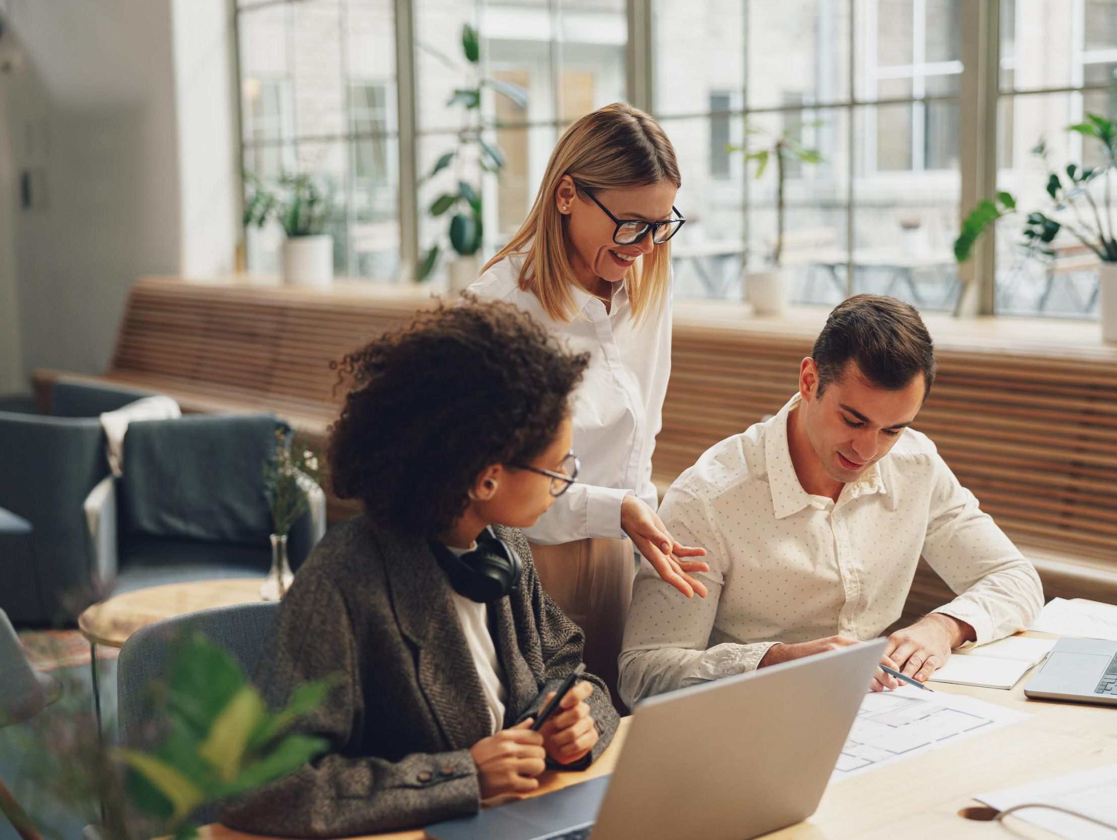 Tre kollegaer samarbeider rundt et bord i et lyst kontor med store vinduer og grønne planter.