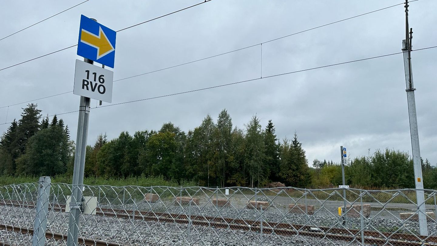 Jernbaneskilt med blå pil og tekst 116 RVO ved dobbeltspor og skog i bakgrunnen